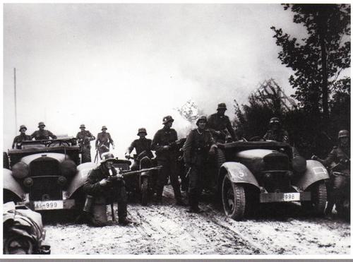 Officers and enlisted men of the Leibstandarte SS Adolph Hitler take a break on a dusty Polish road as a building burns behind t.jpg